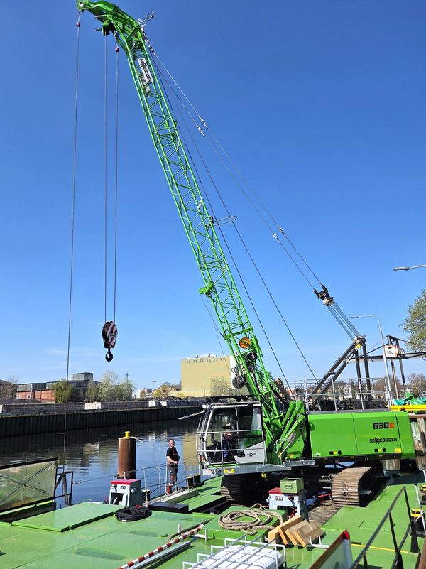 A SENNEBOGEN 630 E cable excavator mounted on a pontoon, being used as a floating grab on a waterway.