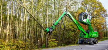 A SENNEBOGEN 718 E felling excavator equipped with a grapple saw performing tree maintenance work along a wooded road.