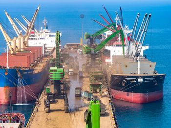 SENNEBOGEN 895 E Umschlagbagger bei der Schiffsabfertigung am Hafenpier von Tosyalı mit Frachtschiffen und wartenden Lkw.
